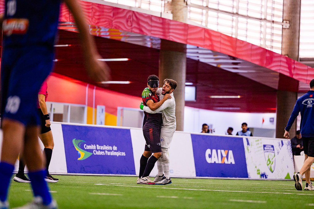 Dois jogadores de futebol de cegos, Matheus e Raynã, da APACE, se abraçando em campo sintético coberto. Um deles está com uniforme preto e vermelho, meião preto e chuteira clara; o outro jogador, de uniforme todo branco, o envolve num abraço apertado, com expressão de alegria na comemoração de gol. Ao fundo há placas de publicidade nas laterais do campo; em uma delas está escrito “Comitê Brasileiro de Clubes Paralímpicos” com o logotipo em verde, amarelo e azul, e mais à direita outra placa com o logotipo da “CAIXA”
