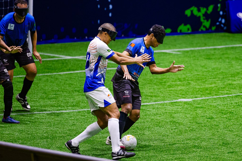 Raynã, da APACE, de camisa azul e short preto, protege a bola com o corpo enquanto Carlinhos, da ADESUL, de uniforme branco com detalhes azuis e short branco, tenta alcançá-la. Os dois atletas estão com vendas nos olhos e disputam a posse de bola em campo sintético. Ao fundo, outro jogador da APACE acompanha a jogada